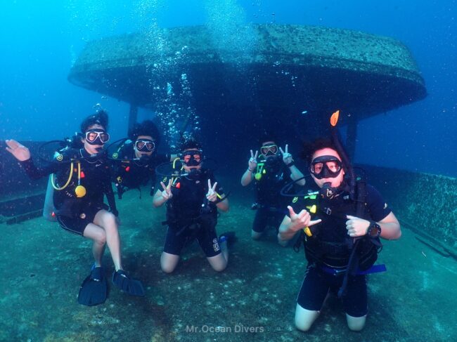 水中にある沈船を背景に5人のダイバーが写っています