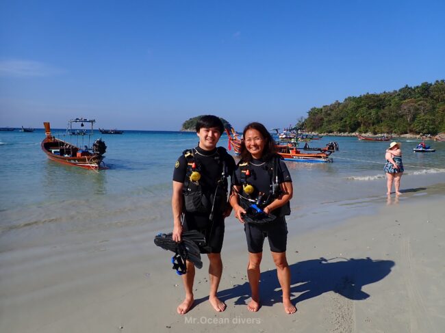 ビーチの上で海を背景にダイビング器材を背負ったダイバーが2人見えます。