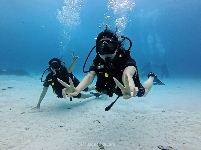 白い砂地と青い海の中で2人のダイバーがピースをしているのが見えます